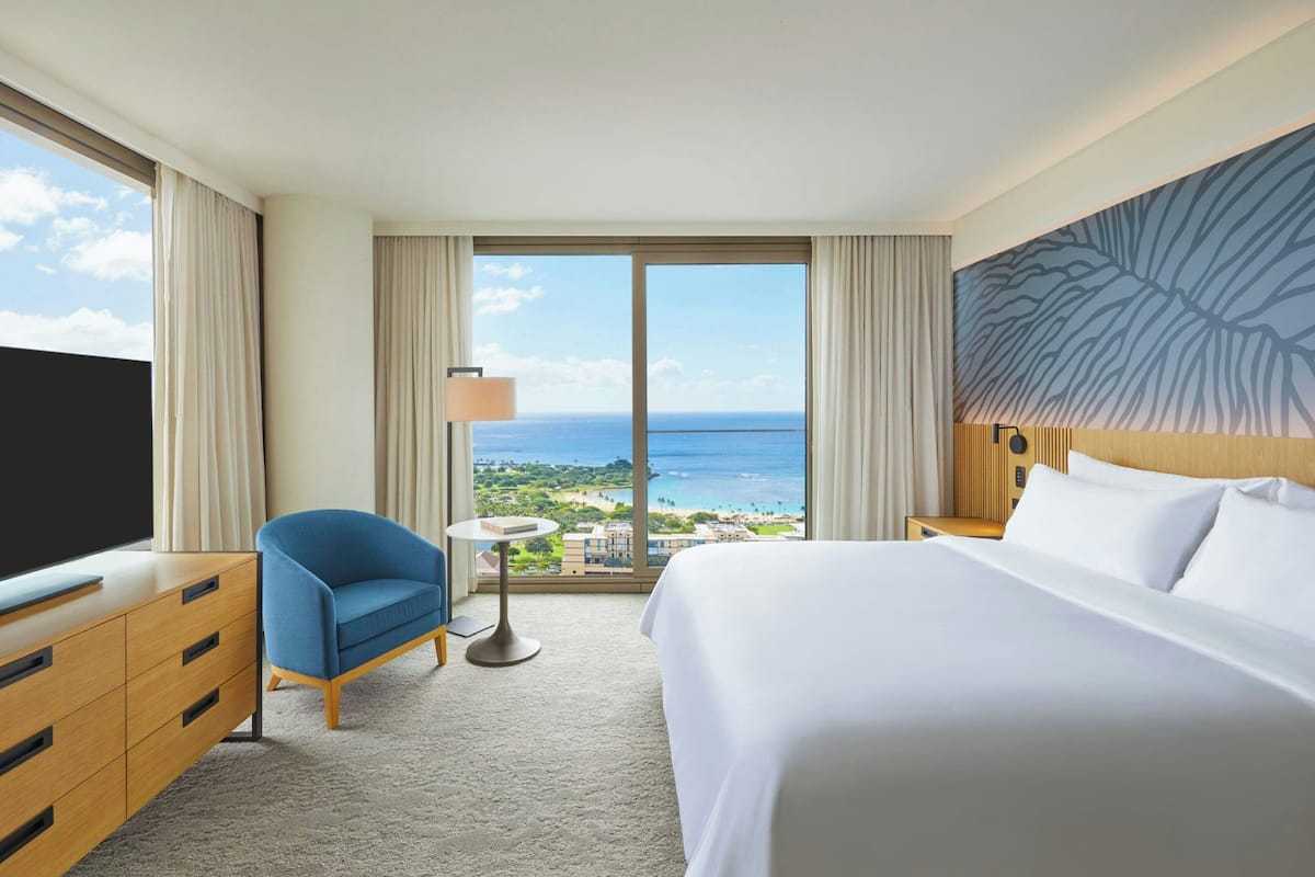 A hotel room with a large bed, a TV, a desk, a chair, and a view of the ocean.