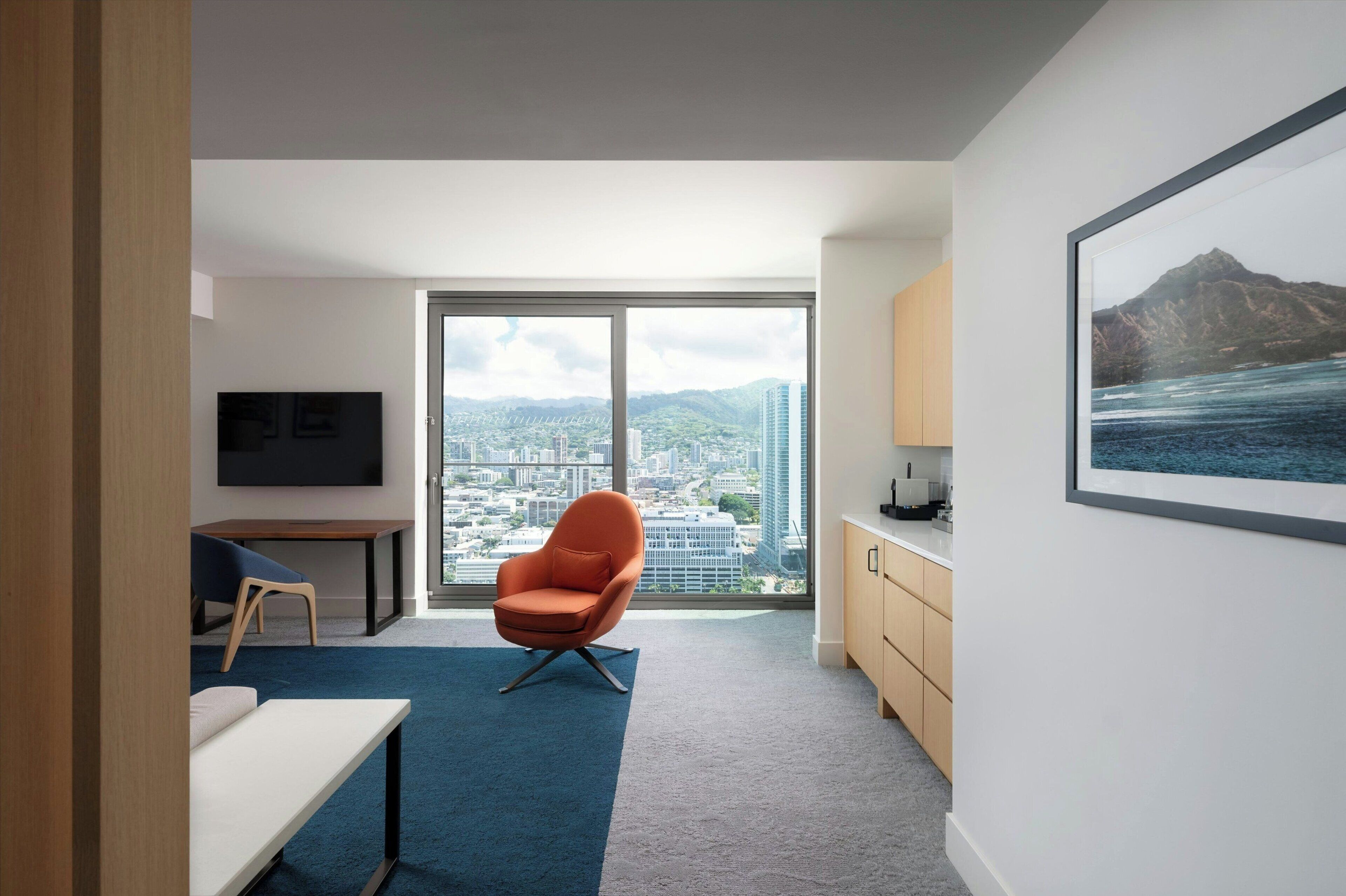 Una habitación de hotel moderna con un amplio ventanal que ofrece vistas panorámicas de la ciudad, un sillón naranja confortable, un televisor de pantalla plana y un diseño minimalista.
