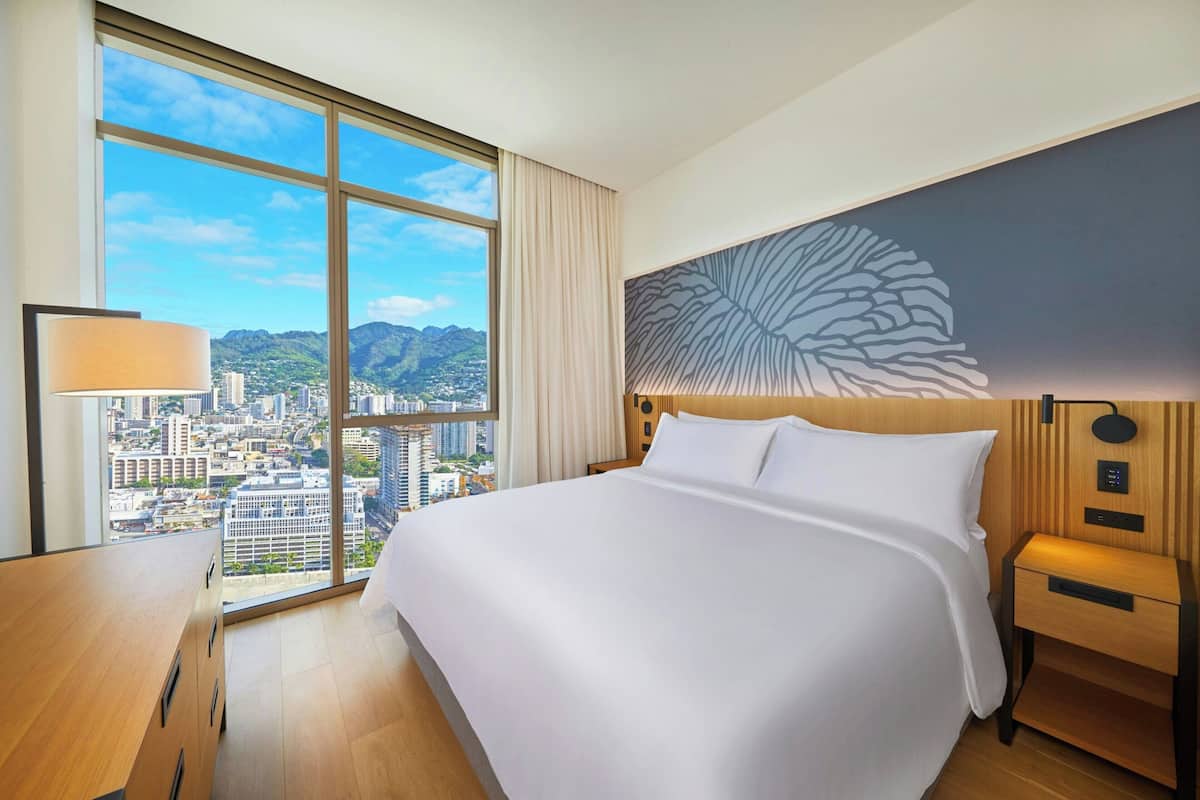A hotel room with a large bed, a desk, a nightstand, and a view of a cityscape through a large window.