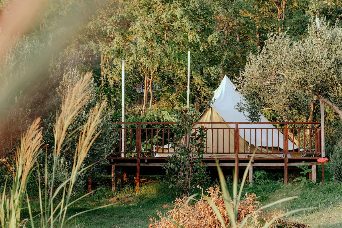 Une terrasse en bois surmontée d’une structure blanche en forme de tente, entourée d’une végétation luxuriante et d’herbes hautes.