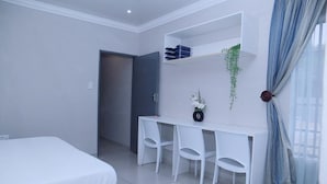 A bedroom with a bed, a desk with chairs, a small plant, and a window with curtains.