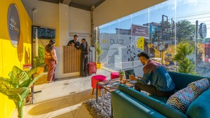 Un espace de travail moderne doté d’un mur en verre, d’un mur d’accent jaune et d’un coin salon avec un ordinateur portable et une plante.