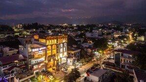 Un immeuble à plusieurs étages avec une enseigne lumineuse, une rue au premier plan et un paysage montagneux en arrière-plan.