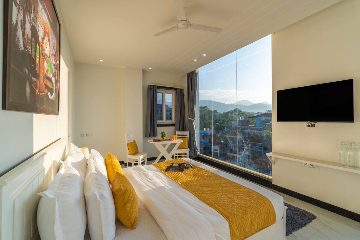 Ein Hotelzimmer mit Bett, Schreibtisch, Stuhl, Fernseher und einem großen Fenster mit Blick auf die Stadt.