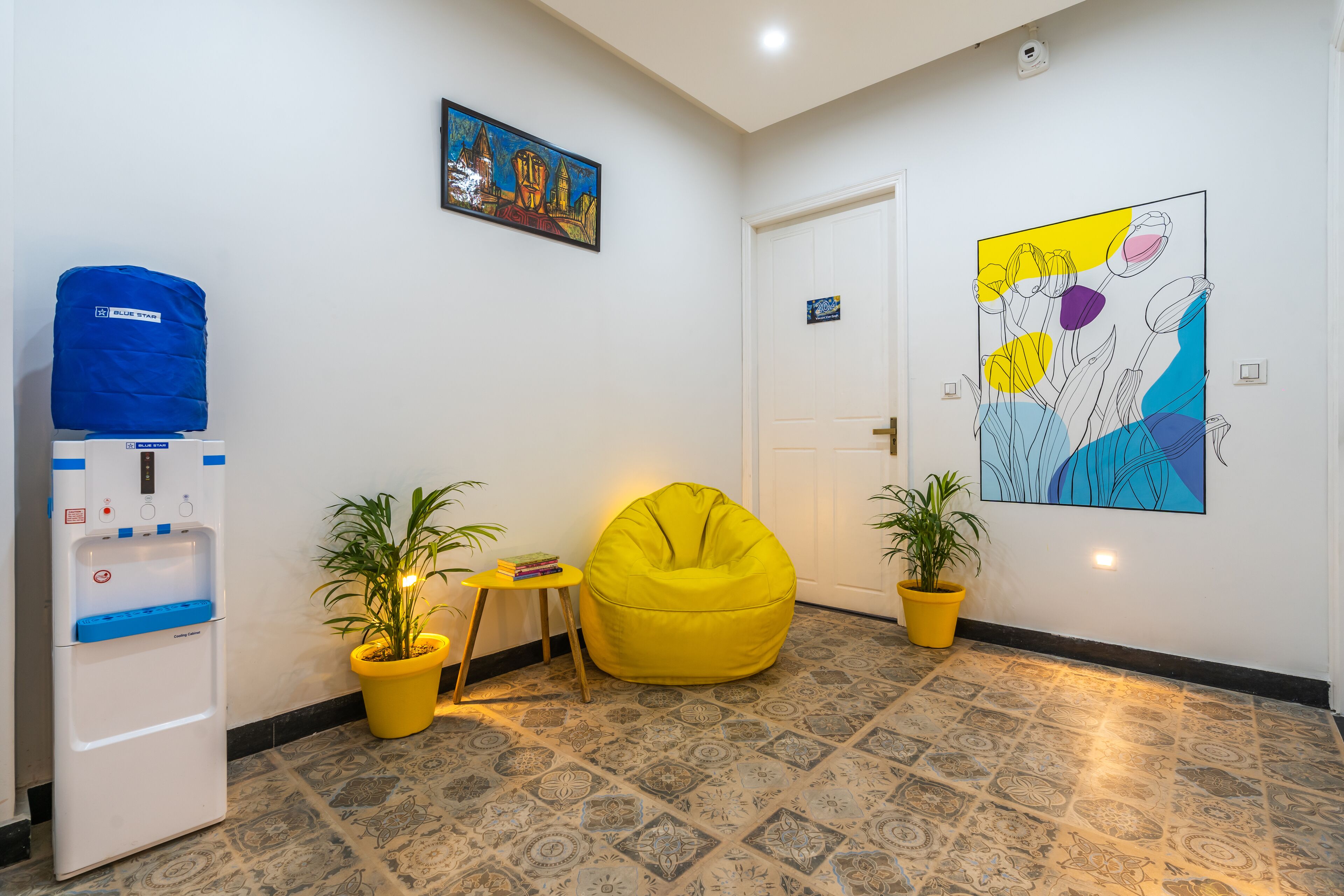 Een kamer met een waterdispenser, een gele relaxfauteuil, twee potplanten en een kleurrijk abstract schilderij aan de muur.