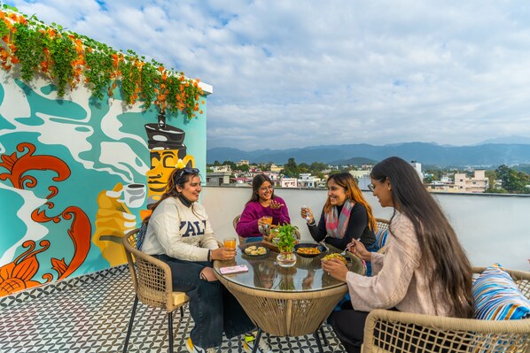 Um grupo de pessoas jantando em uma mesa ao ar livre, com vista para montanhas e uma paisagem urbana ao fundo.