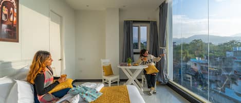 A modern hotel room with a large bed, a small table, and a view of the city through large windows.