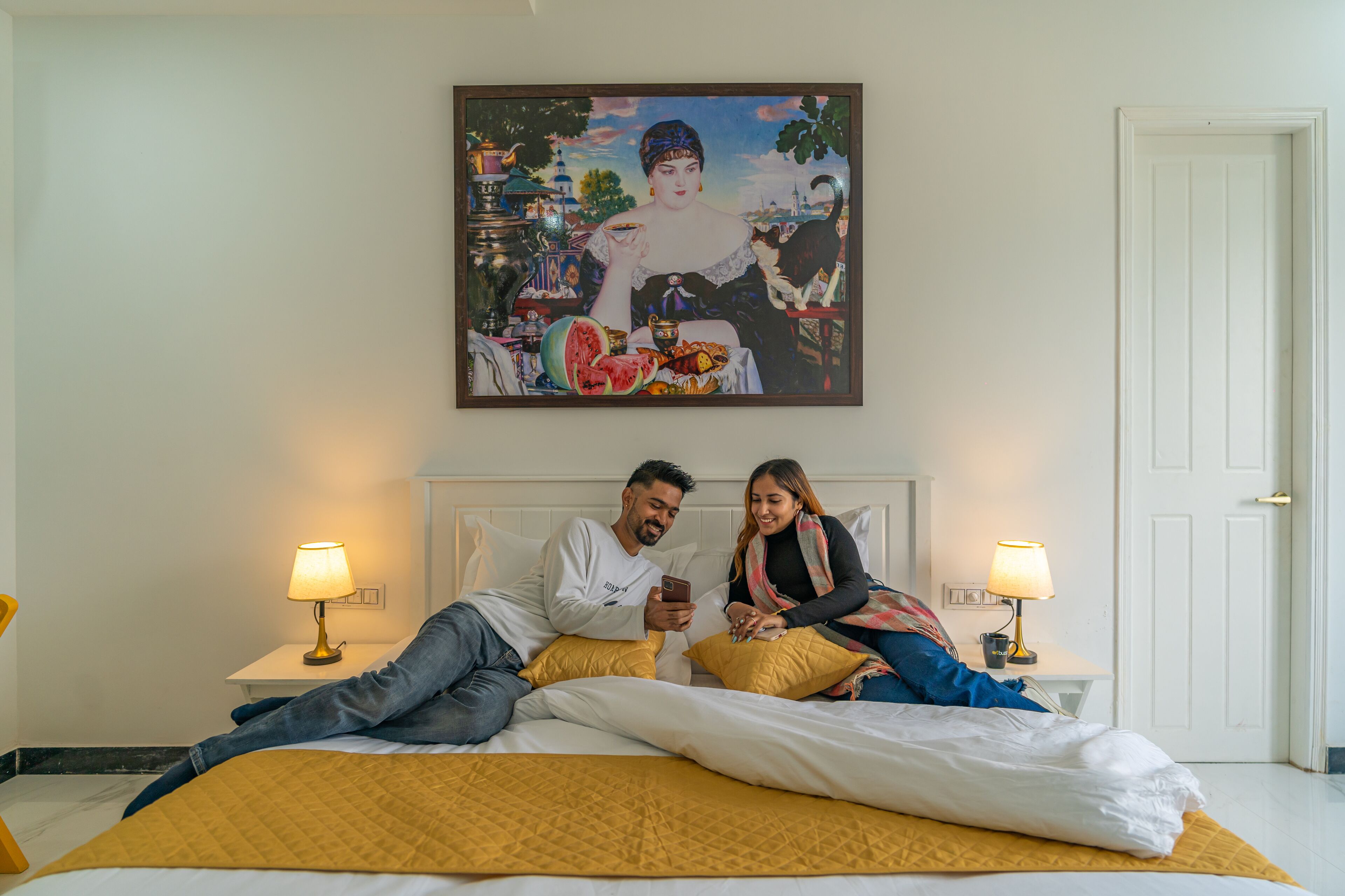 Una pareja en una cama con un cuadro encima, dos lámparas de noche y una puerta blanca.