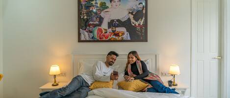 A couple on a bed with a painting above them, two bedside lamps, and a white door.