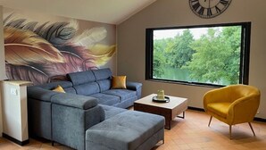 A living room with a sectional sofa, a yellow armchair, a coffee table, a clock, and a large window with a view of trees.