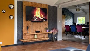 A modern living room with a large flat-screen TV mounted on a wooden wall, a fireplace below, and a dining area visible through an open doorway.