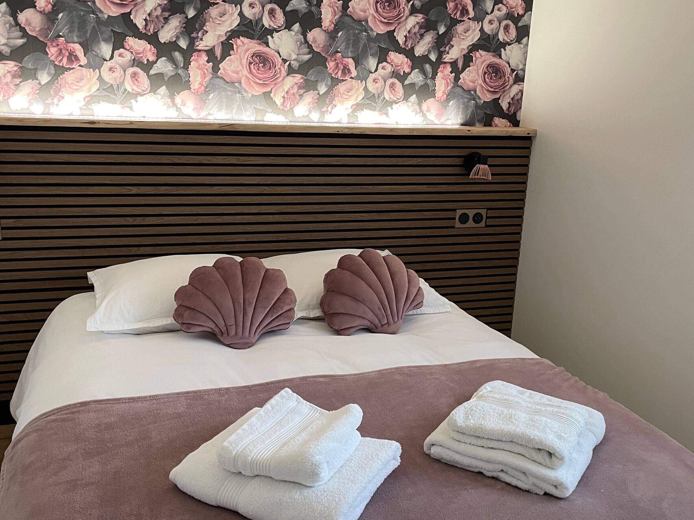 A neatly made bed with pink and white pillows, a wooden headboard, and a floral wallpaper.