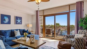 A spacious living room with a blue sofa, a coffee table, and a patterned armchair. A large sliding glass door leads to an outdoor balcony with a view of palm trees and a pool.
