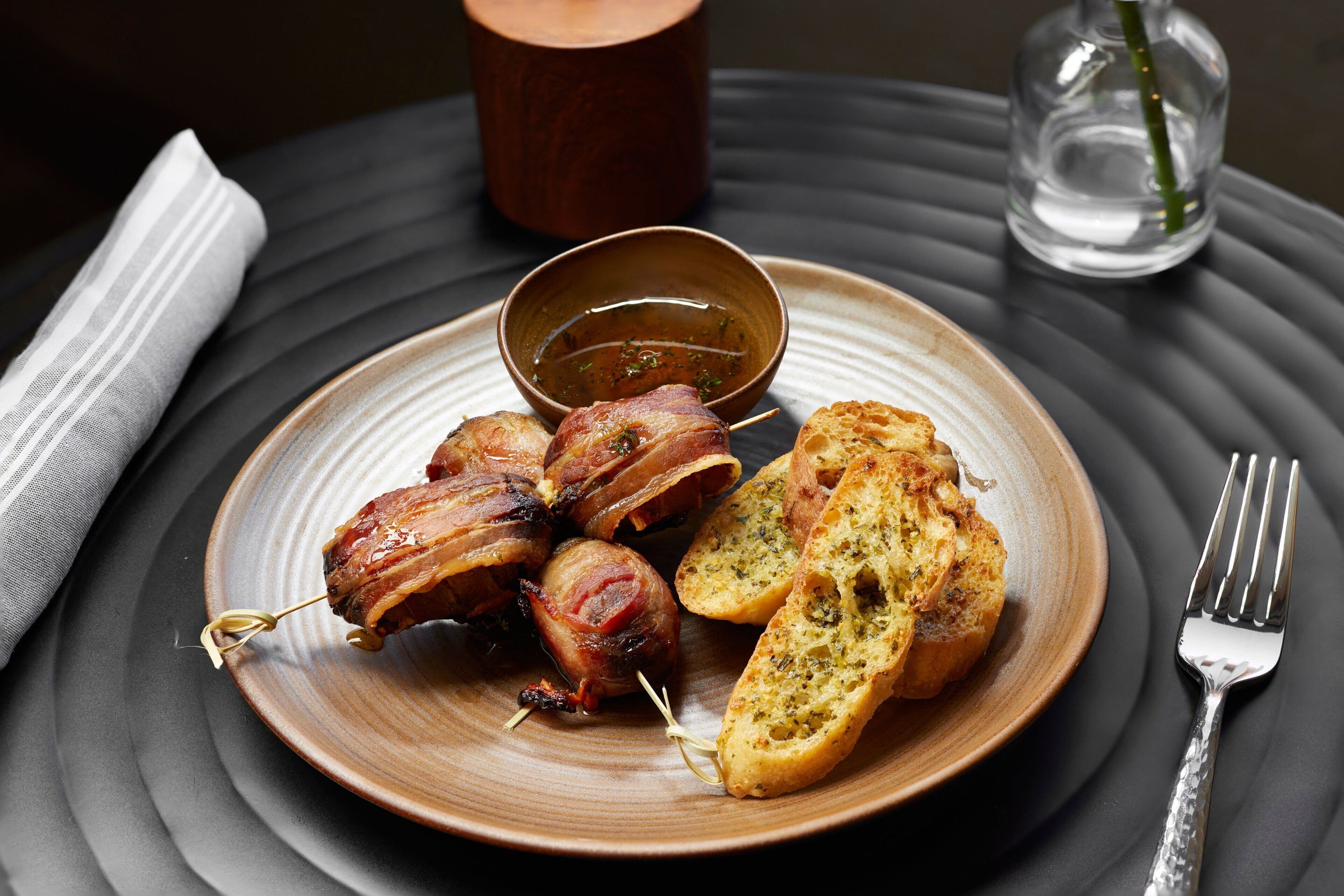 Une assiette contenant des boulettes de viande enroulées de lardons, du pain à l’ail et un petit bol de sauce, le tout présenté sur une assiette foncée.