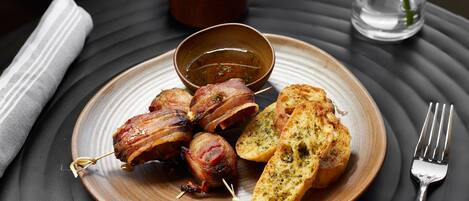 Une assiette contenant des boulettes de viande enroulées de lardons, du pain à l’ail et un petit bol de sauce, le tout présenté sur une assiette foncée.