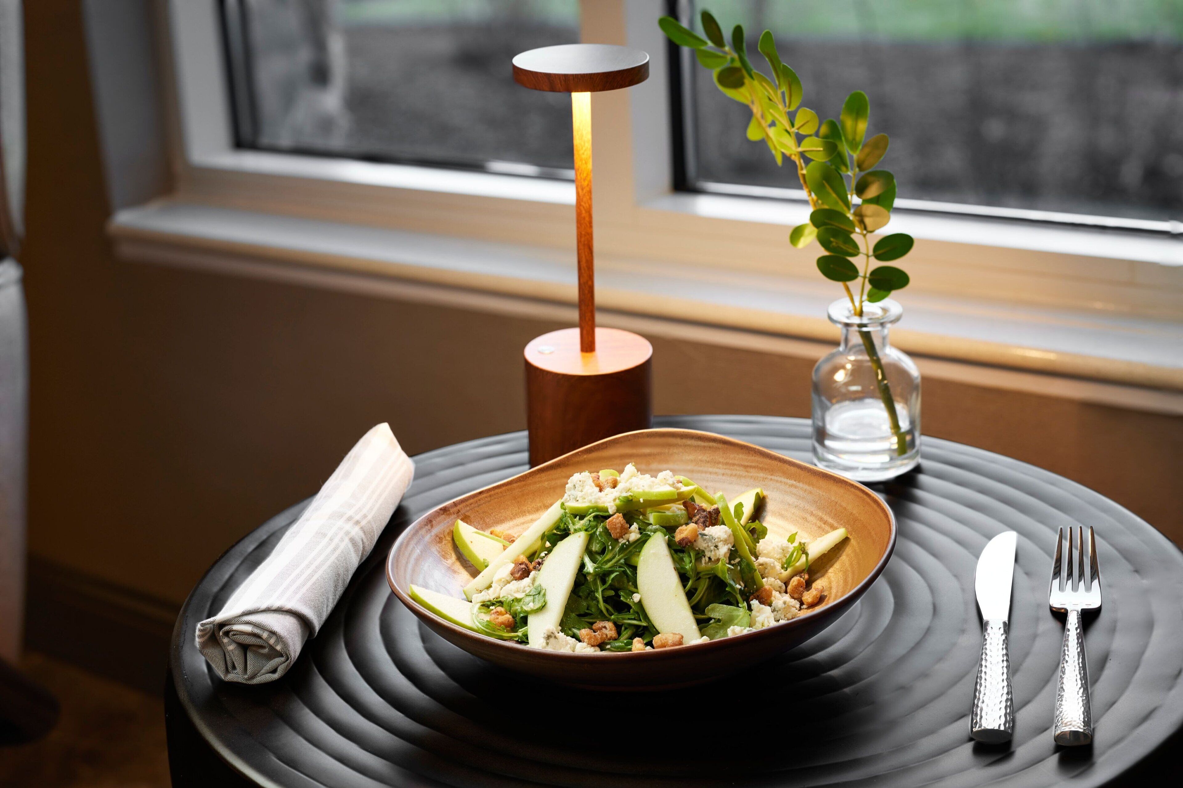 Ein Esstisch mit einer Speisenplatte, einem Servietttuch, Besteck, einer Vase mit einer Pflanze und einer Tischlampe.
