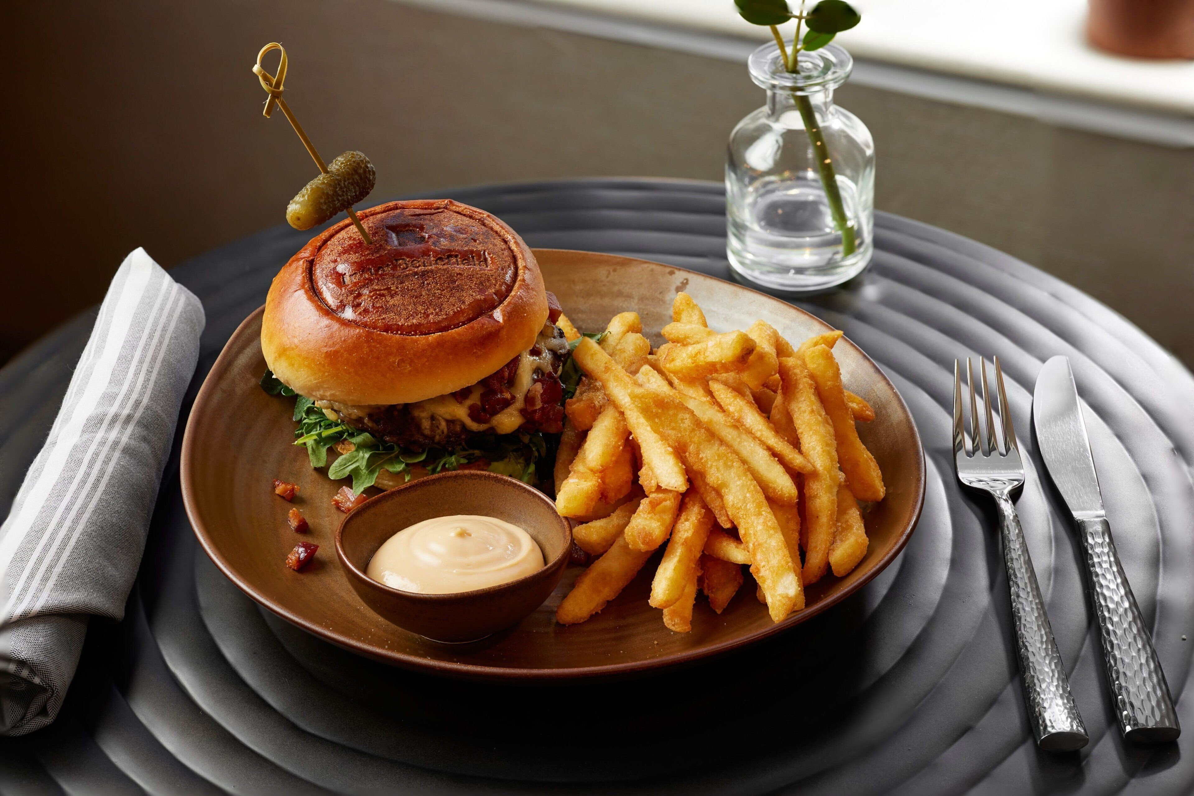 A plate with a burger, fries, and a small bowl of sauce, set on a table with a napkin and cutlery.