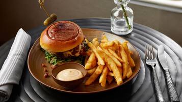 A plate with a burger, fries, and a small bowl of sauce, set on a table with a napkin and cutlery.