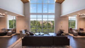 Uma sala de estar espaçosa com uma grande janela que oferece vista para áreas verdes e edifícios, teto de madeira alto e móveis modernos.