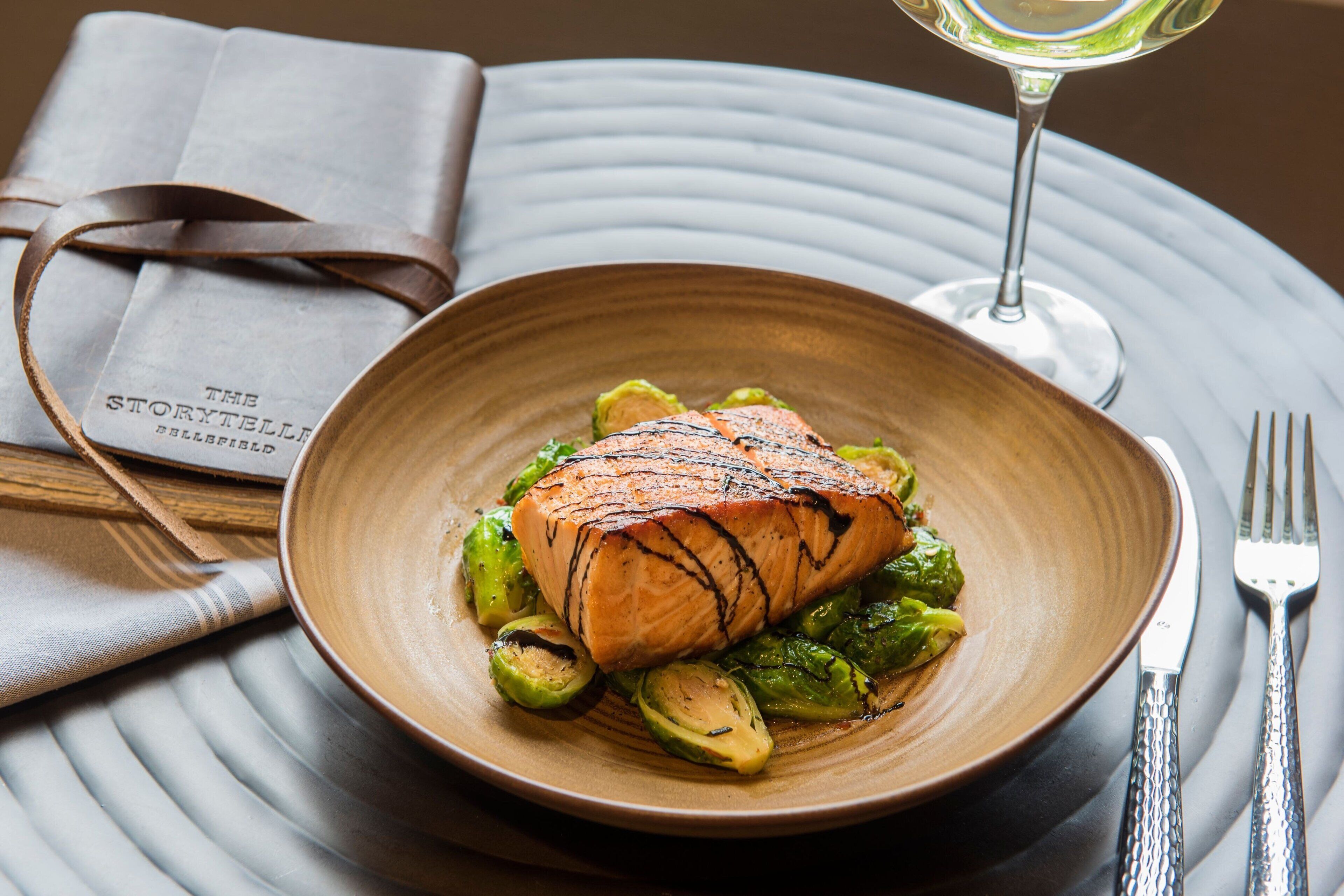 Ein Teller gegrillter Lachs mit Rosenkohl auf einer strukturierten Platte, dazu ein Glas Weißwein und eine gefaltete Speisekarte.