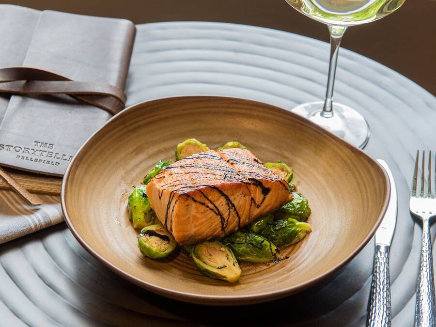 Ein Teller gegrillter Lachs mit Rosenkohl auf einer strukturierten Platte, dazu ein Glas Weißwein und eine gefaltete Speisekarte.