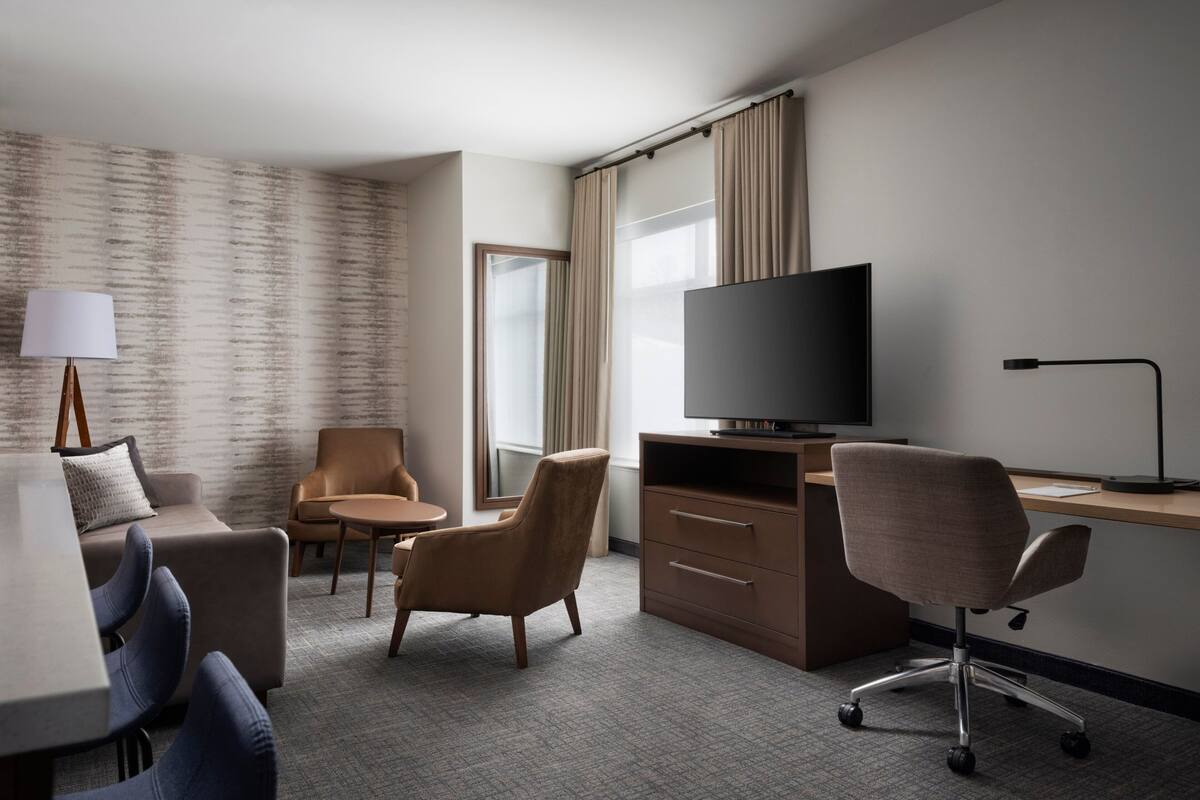 Une chambre d’hôtel avec un canapé, deux fauteuils, un bureau avec une télévision et une lampe.