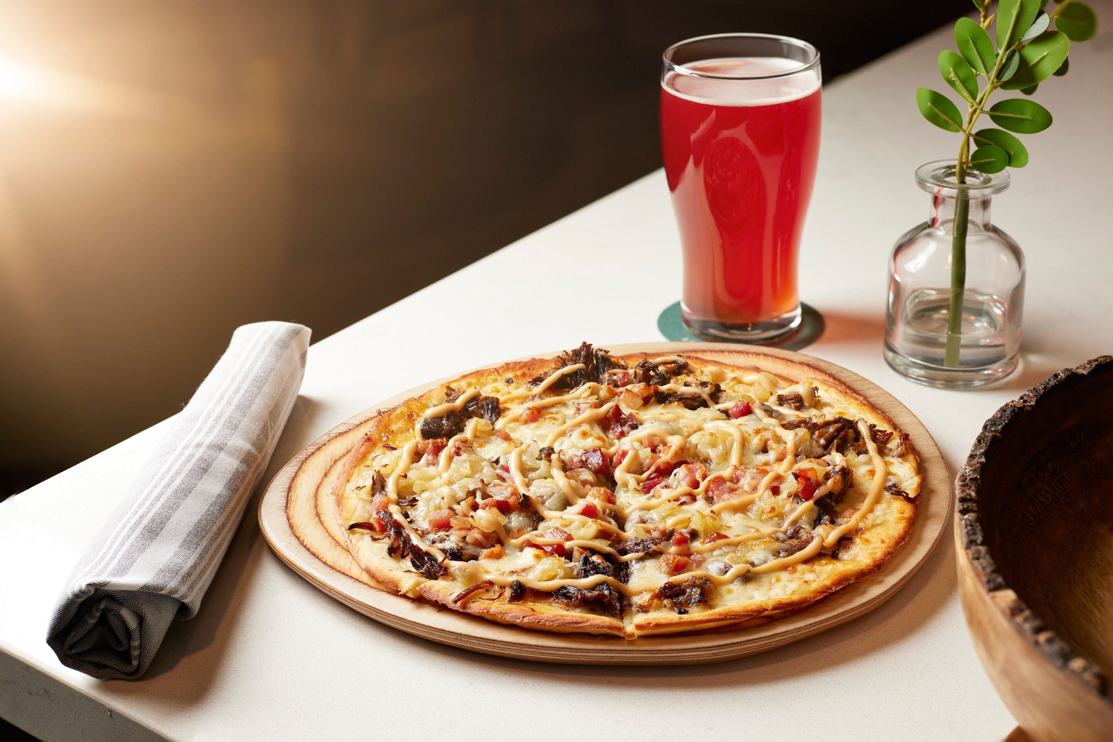 A pizza with various toppings on a wooden board, a glass of red beverage, a small vase with a plant, and a rolled-up napkin on a table.