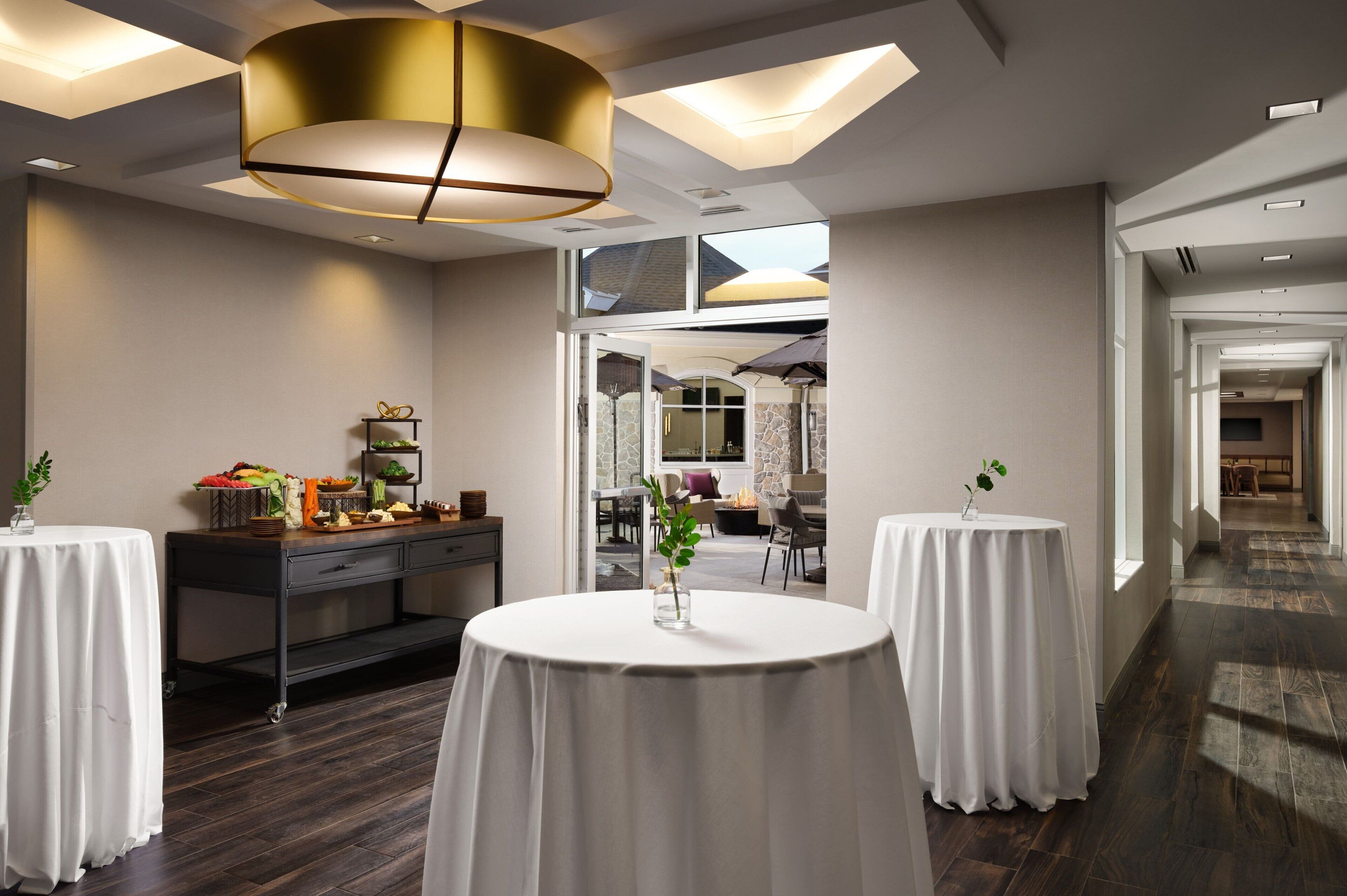 A modern hotel lobby with two round tables, a central light fixture, and a view into a dining area.