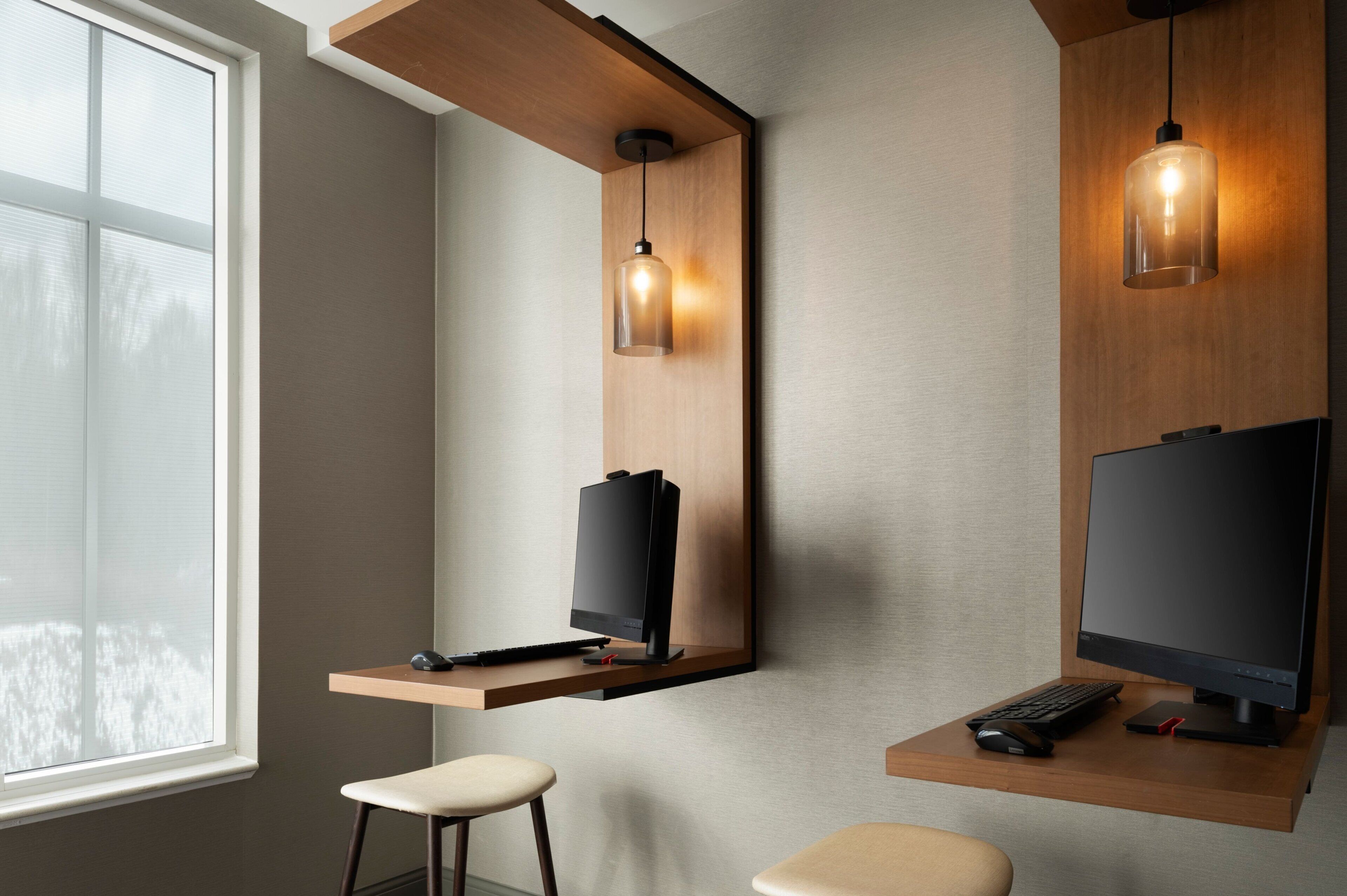 A modern office space with two desks, each with a computer monitor, keyboard, and mouse. There are two pendant lights hanging from the ceiling, and a window providing natural light.