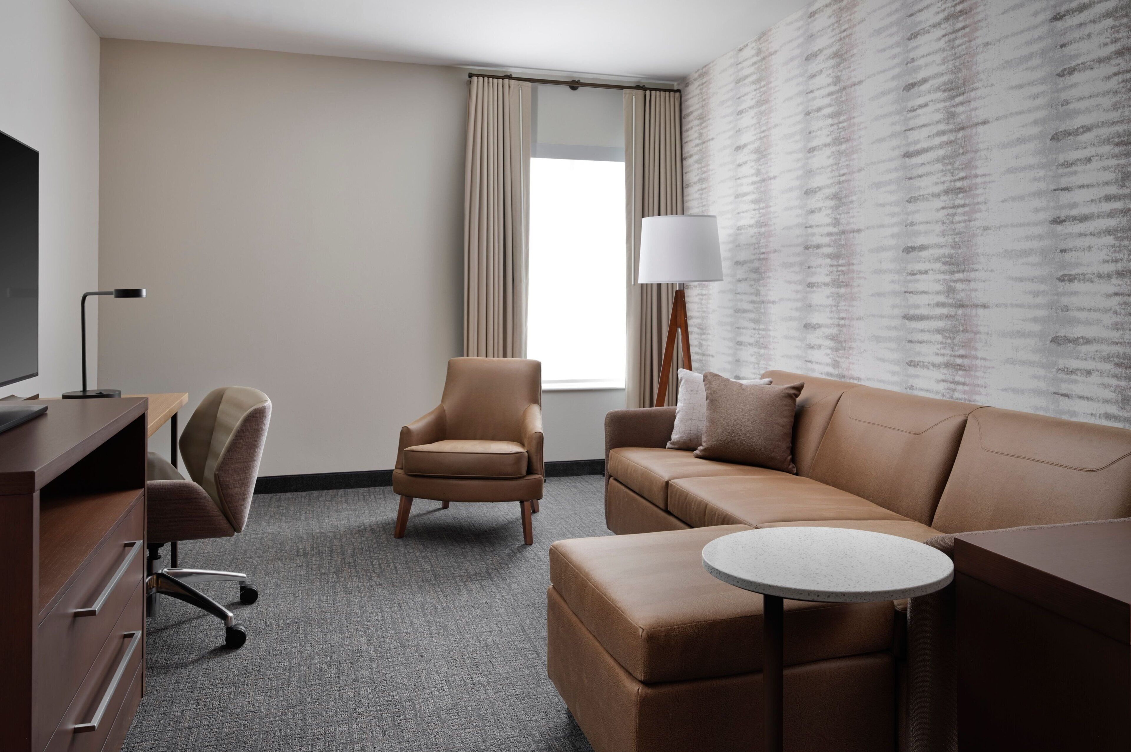 A hotel room with a brown leather sofa, a brown leather armchair, a small round table, a TV on a wooden desk, and a window with curtains.