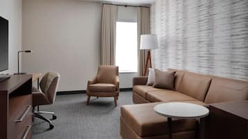 A hotel room with a brown leather sofa, a brown leather armchair, a small round table, a TV on a wooden desk, and a window with curtains.