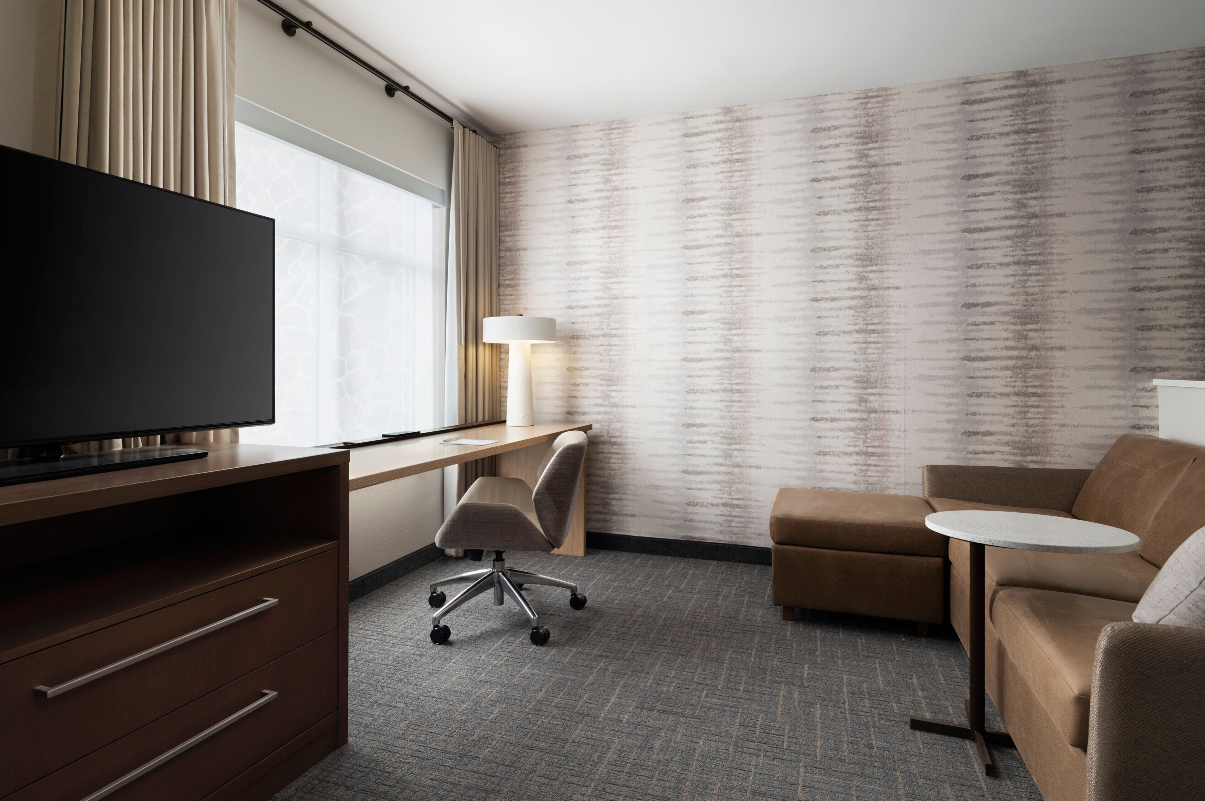 A hotel room with a flat-screen TV, a desk with a lamp, a sofa, and a small round table.