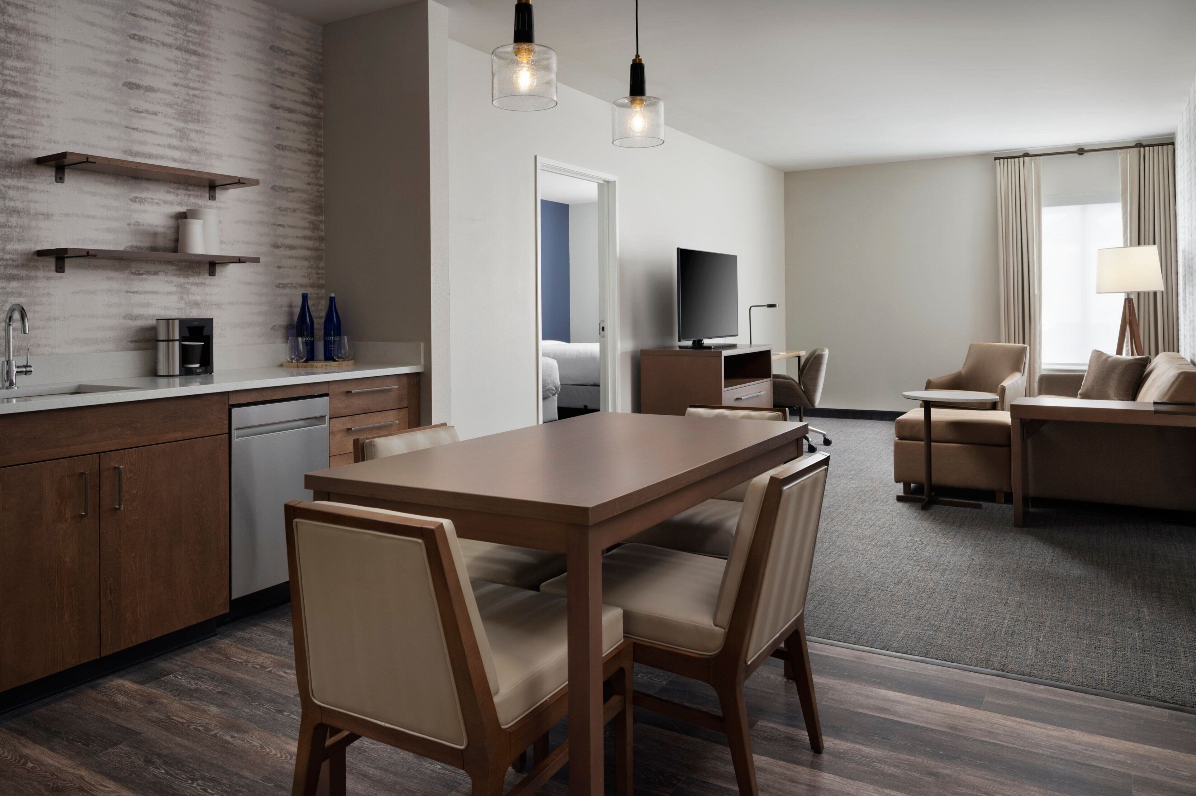 A modern hotel room with a wooden dining table, beige chairs, a flat-screen TV, a sofa, and a lamp.