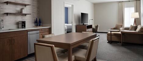 Una habitación de hotel moderna con una mesa de comedor de madera, sillas color beige, un televisor de pantalla plana, un sofá y una lámpara.