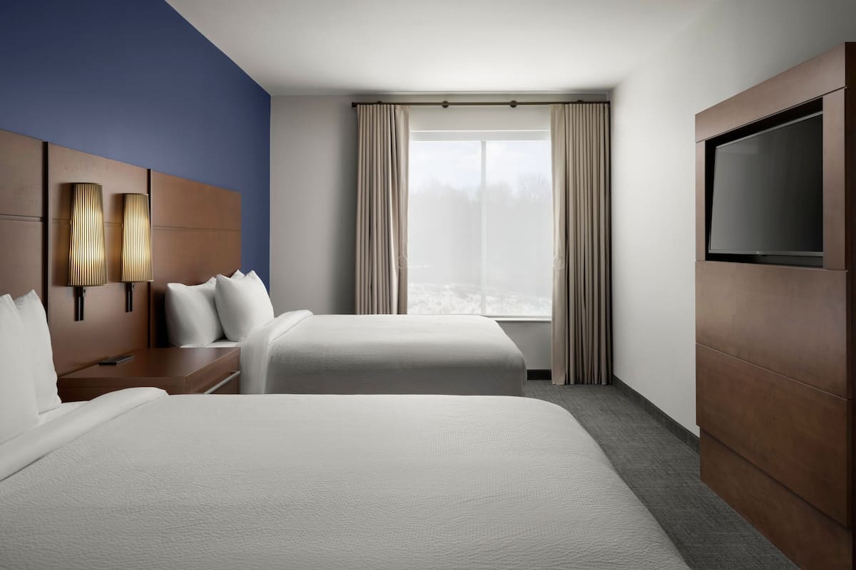 Une chambre d’hôtel avec deux lits, une télévision, une fenêtre avec des rideaux et un luminaire fixé au mur.