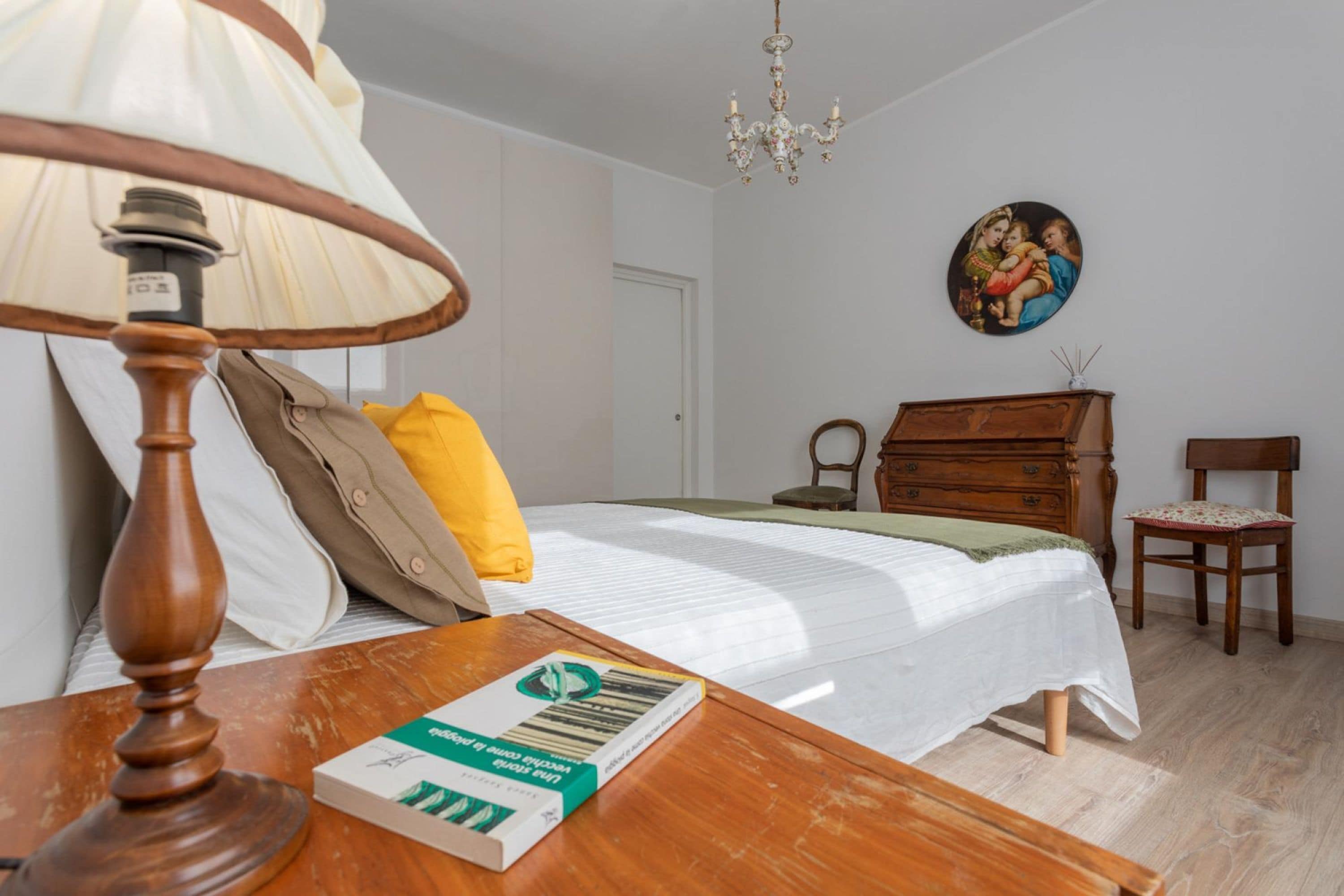 Une chambre à coucher bien rangée, avec un lit, une table de chevet sur laquelle se trouve un livre, une lampe, une chaise et un tableau accroché au mur.