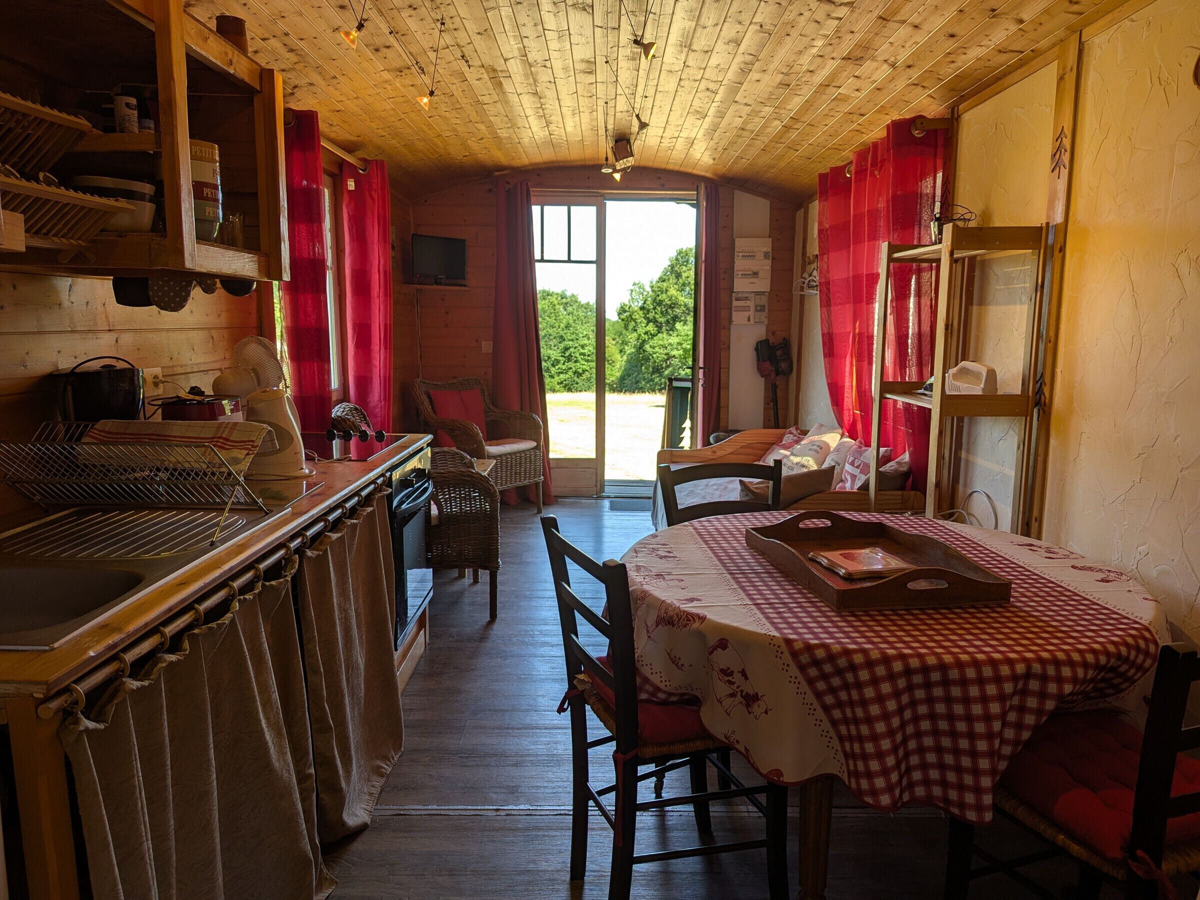 Un intérieur rustique en bois, avec une table à manger, des chaises, un coin cuisine doté d’étagères et une vue sur l’extérieur, visible à travers une porte.