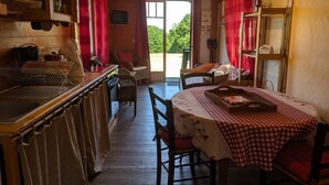 A rustic wooden interior with a dining table, chairs, a kitchen area with shelves, and a view through a doorway.