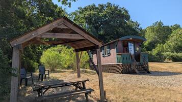 Une structure rustique en bois, avec une table de pique-nique et des chaises sous un abri, une petite cabane dotée d’une véranda et un chemin de gravier.