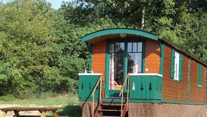 A wooden cabin with a green exterior, a wooden staircase leading to the door, and a picnic table in front.