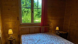 A wooden room with a bed, two bedside lamps, a window with a view of greenery, and a small table with a lamp.