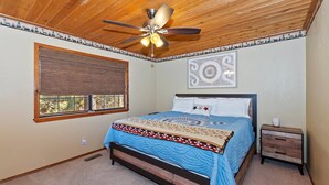 A bedroom with a bed, a ceiling fan, a window with blinds, a nightstand, and a wall art piece.