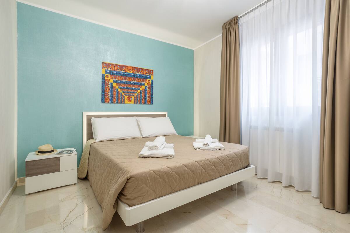 A neatly made bed with a beige bedspread, a white pillow, and a folded towel on the side. A colorful artwork is hung on the teal wall above the bed.