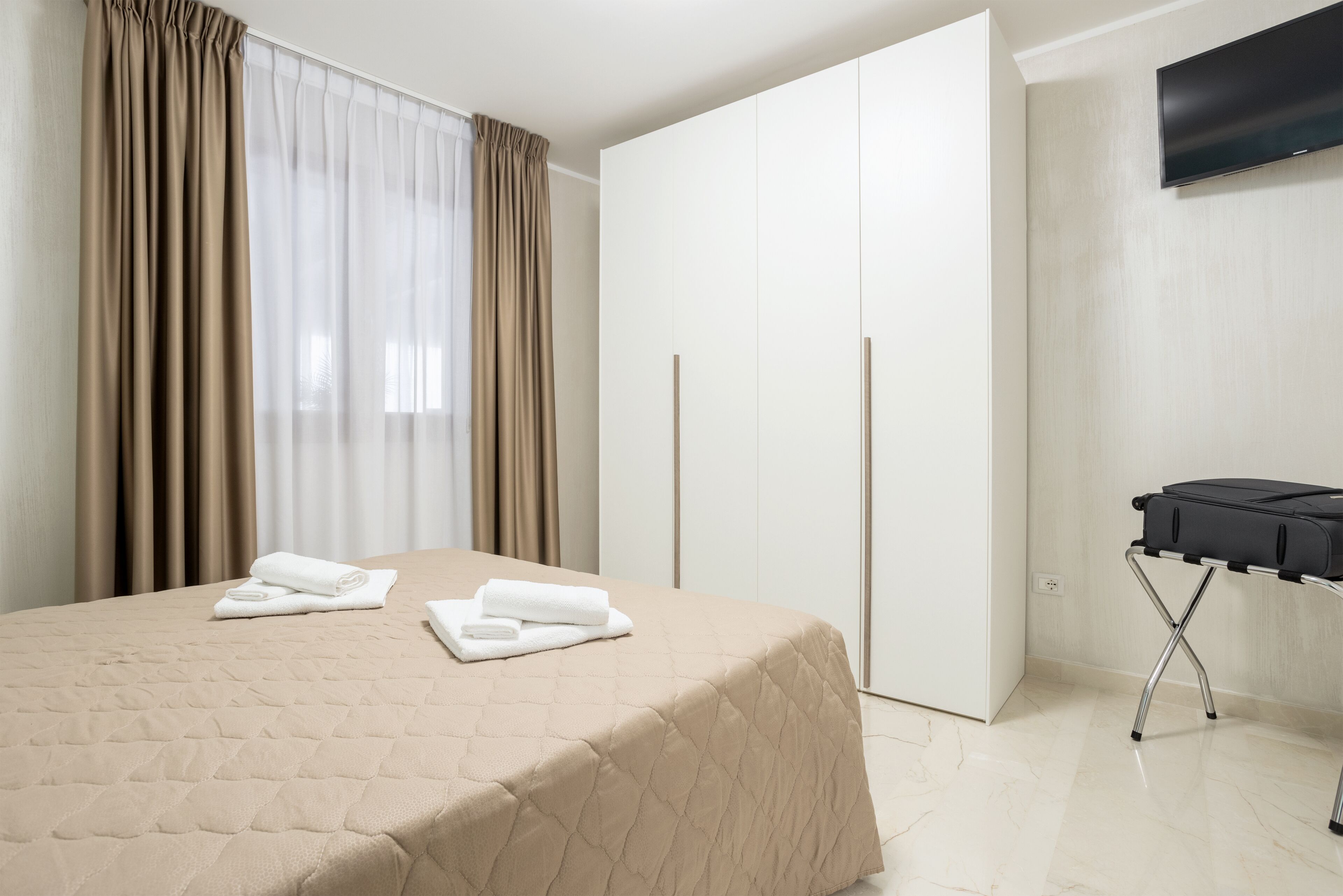 A hotel room with a bed, a wardrobe, a flat-screen TV, and a suitcase on a trolley.
