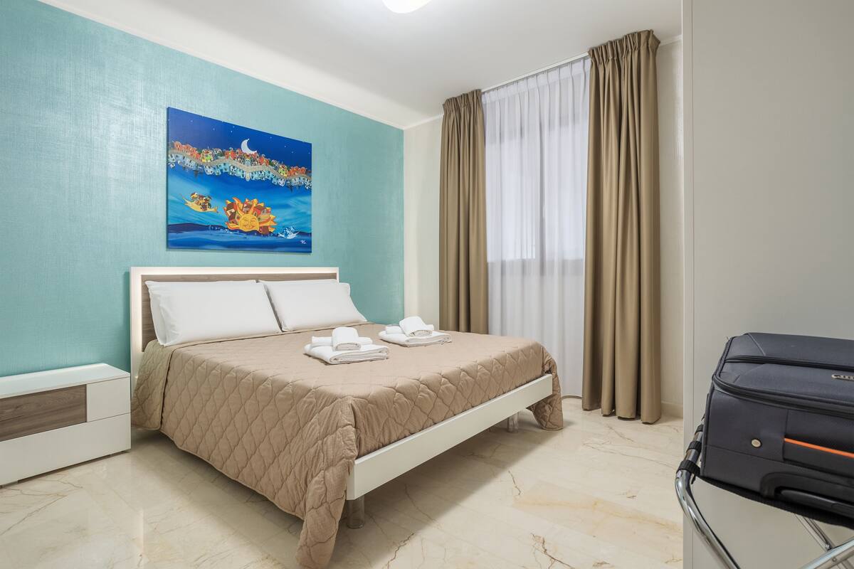 A hotel room with a bed, a painting, a window with curtains, and a suitcase.