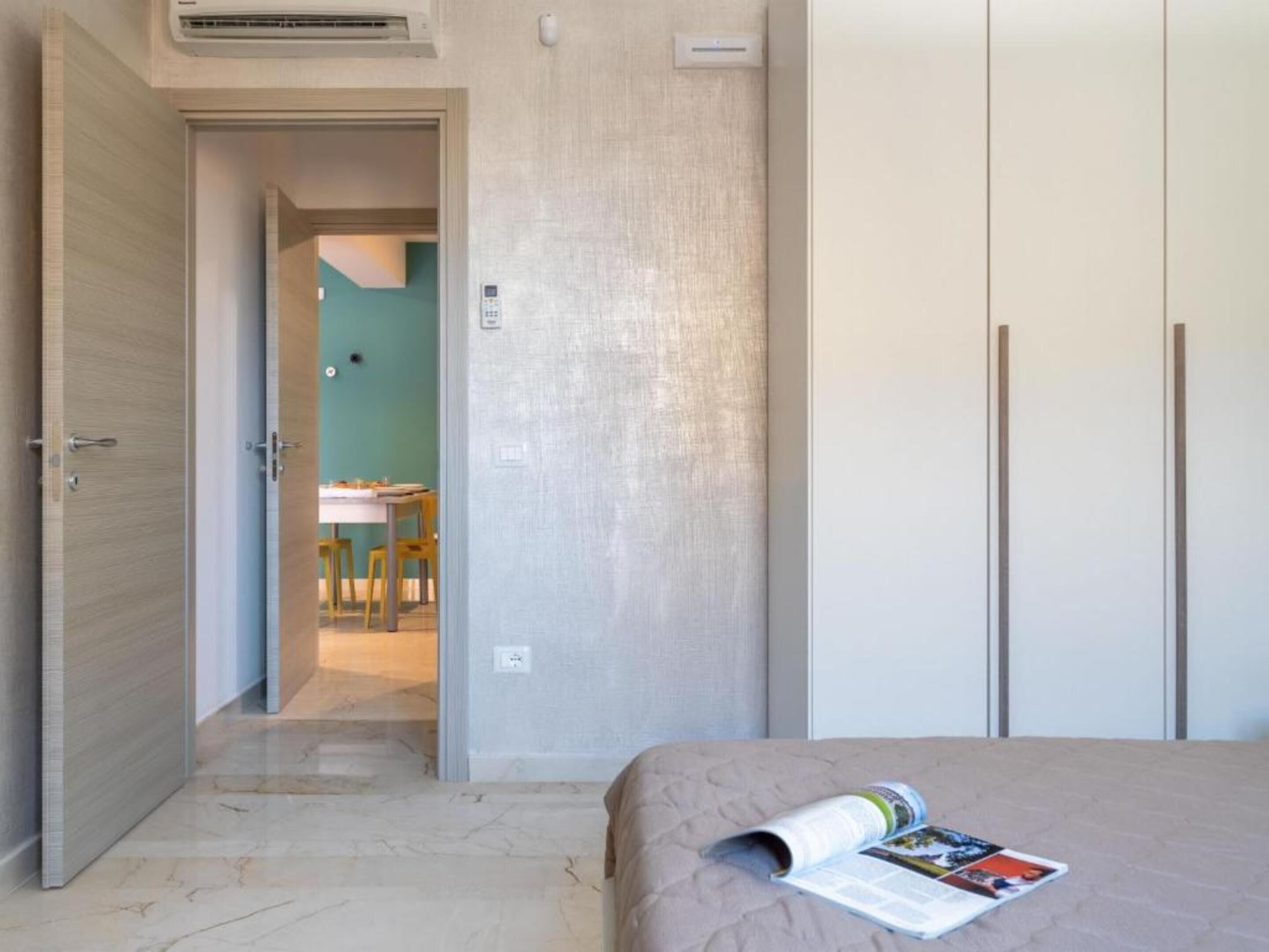 A hotel room with a bed, a magazine on the bed, a door leading to a dining area, and a wall-mounted air conditioner.