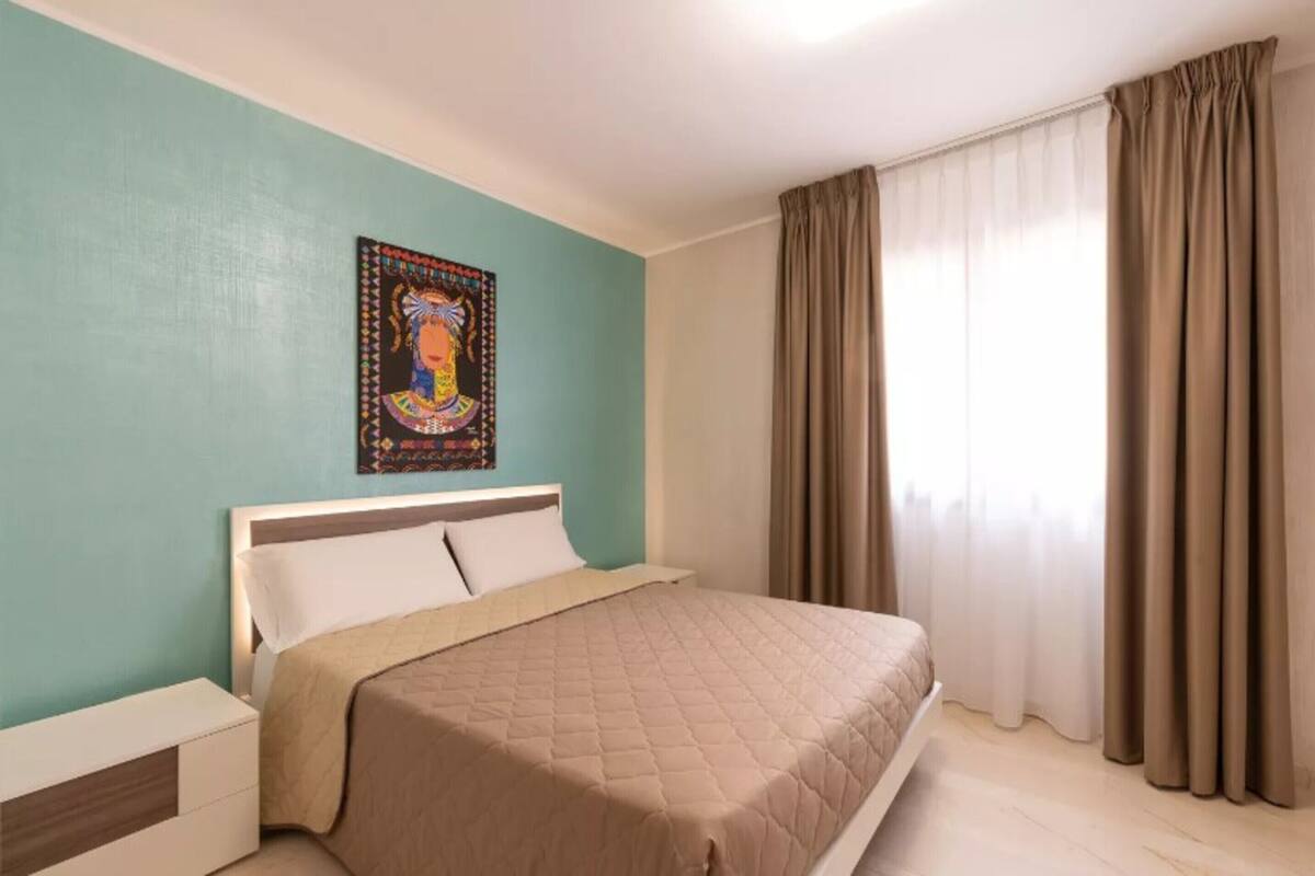 A bedroom with a bed, a nightstand, a wall-mounted artwork, and a window with curtains.