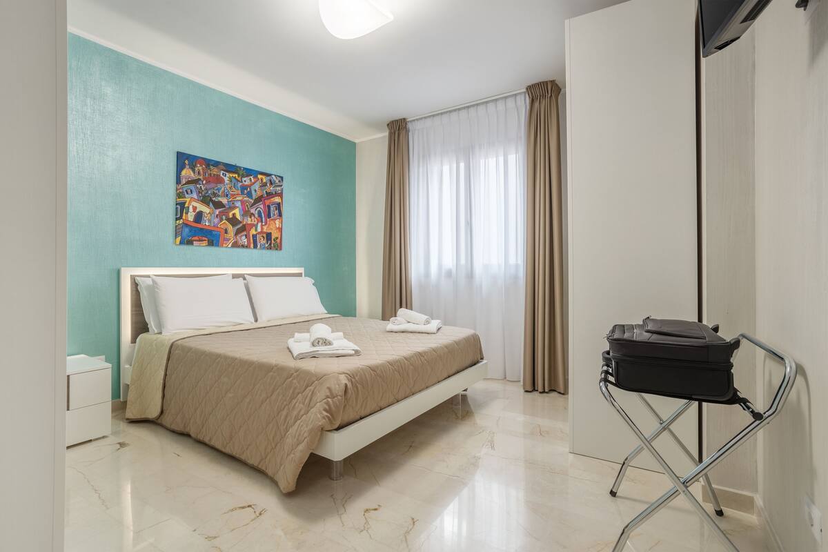 A bedroom with a bed, a painting on the wall, a window with curtains, and a suitcase on a stand.