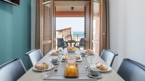 A dining table set with breakfast items, a pitcher of orange juice, and a view of the sea through an open door. - Azzurra Apartments (Balestrate)