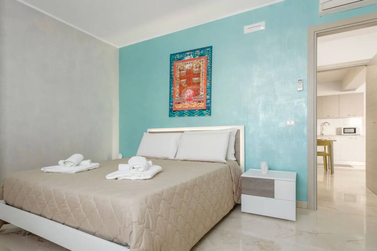A modern hotel room with a bed, a nightstand, a wall-mounted artwork, and a view into the kitchen.